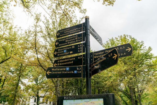 Wegwijzer op het Lange Voorhout in Den Haag met richtingaanwijzingen naar diverse musea, theaters en wandelroutes, omgeven door groene bomen.